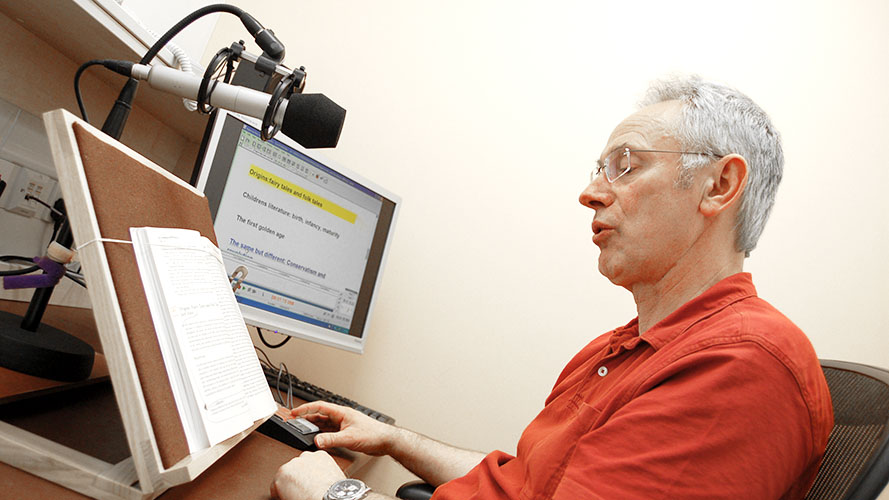 A man reading a book out loud into a microphone, with Publisher on the computer screen.